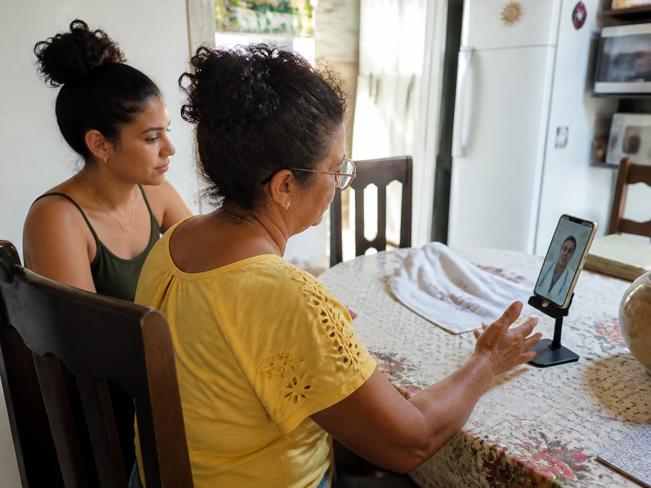 Mulher em atendimento médico por vídeo pelo celular na cozinha de casa, acompanhada pela filha